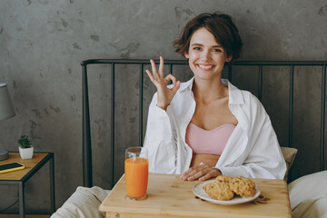Young woman wear white shirt pajama she lying in bed eat breakfast showing ok gesture rest relax...