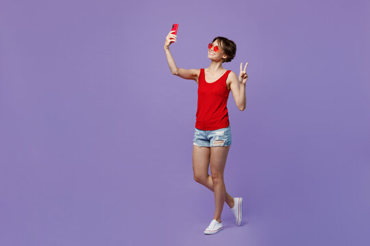 Full Body Young Smiling Woman 20s She Wear Red Tank Shirt Eyeglasses Doing Selfie Shot On Mobile Cell Phone Post Photo On Social Network Show V-sign Isolated On Plain Purple Backround Studio Portrait