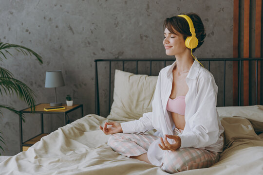 Young woman wear white shirt pajama headphones she lying in bed listen to music mantra meditating hold hands in yoga om gesture rest relax spend time in bedroom lounge home in own room hotel wake up.