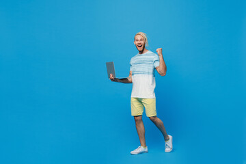 Full size young blond man with dreadlocks 20s he wear white t-shirt hold use work on laptop pc computer do winner gesture isolated on plain pastel light blue background studio People lifestyle concept