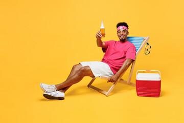 Full size smiling happy cheerful fun young man 20s he wear pink t-shirt bandana near hotel pool raise toast drink beer look camera isolated on plain yellow background. Summer vacation sea rest concept