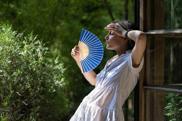 Young woman suffering heatstroke outdoors using fan to get fresh air. Unhappy girl feel bad of hot temperature outside. Frustrated female tired of summer season heat in fatigue touch forehead sweating - obrazy, fototapety, plakaty