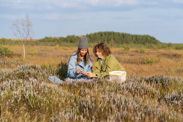 Fototapeta na wymiar Couple of young women best friends sits sorting berries on autumn meadow grass against blurry green forest. Women wearing blue and green jackets enjoy picnic spending time on nature at sunlight