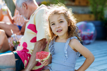 Cute little girl in swimsuit eating watermelon corn and sitting next to pool with family at garden party.