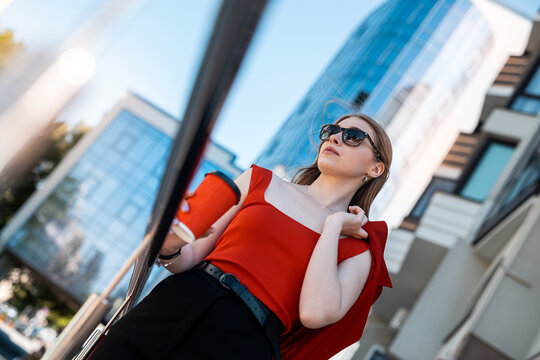 A Young Woman Drinks Coffee On The City Streets Of A Metropolis In Autumn Or Early Spring.