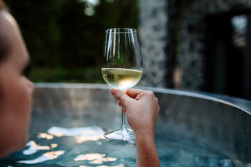 Unrecognizable young woman relaxing with glass of wine in hot tub outdoor in nature.