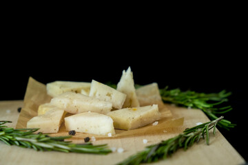 Sliced cheese is laid out on a wooden board with rosemary on a black background