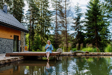 Fototapeta na wymiar Woman, sitting on a pier,near private lake, relaxing, enjoying cup of morning coffee on summer vacation in mountains.