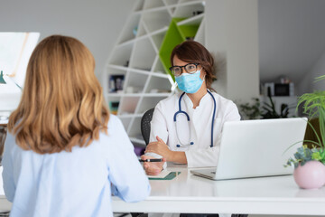Female doctor wearing face mask and consulting her patient while sitting at desk in doctor's office