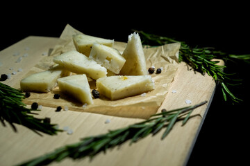 Sliced cheese is laid out on a wooden board with rosemary on a black background