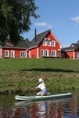 Happy russian tourist child, kid and water sport, active rest, recreation in Fishing village, Ryazan region, Russia. Swimming on SUP-board. Boy and summer water sport, rest with SUP-board. Red houses