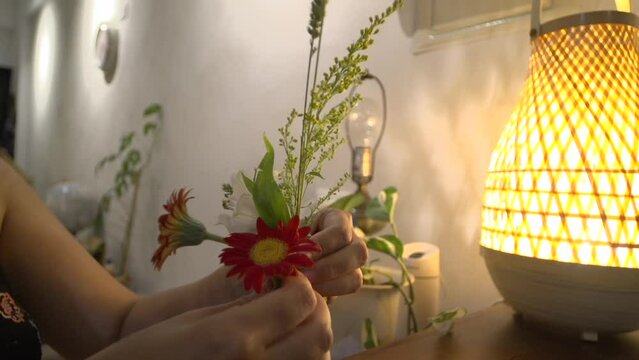 Hands of woman arranging and organizing small bouquet of flowers into glass vase next to bamboo lamp