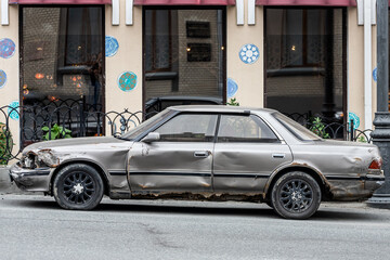An old abandoned  silver car sedan with a broken  doors, broken glass, flat wheels against the background of a city