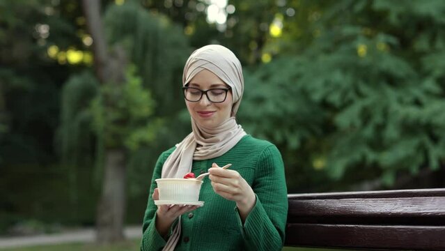Young attractive muslim woman eating salad sitting on the bench on the park. Beautiful young arab female enjoying a fresh salad outdoor.