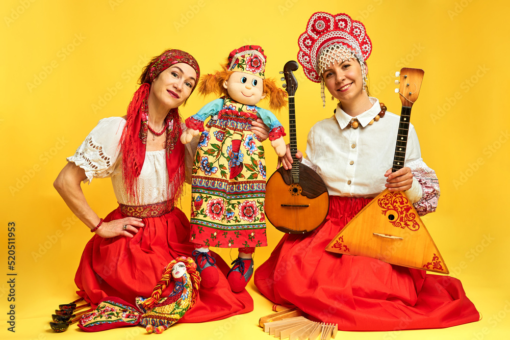 Wall mural Women musicians in Russian folk dresses with musical instruments on a yellow studio background. Happy artists from Russia in national clothes with stringed musical instruments with smiles on faces