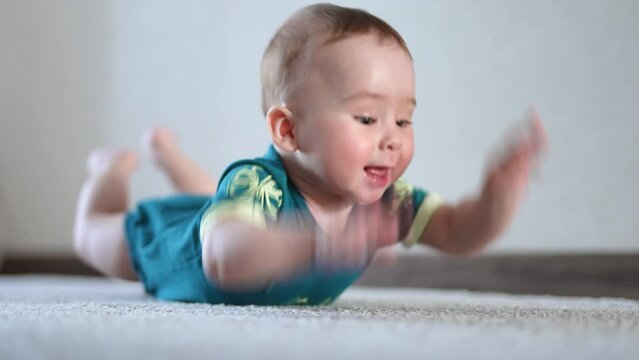 Lovely active baby boy lies on the soft carpet on his belly. Adorable child claps legs and hands by the floor. Light blurred backdrop.
