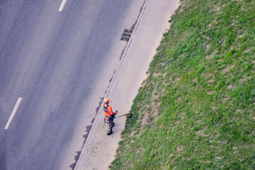 A worker with a lawn mower mows the grass by the road, top view. A man in an orange work uniform...