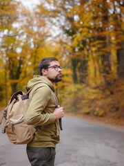 Young handsome man posing in autumn forest on road. young hipster guy with backpack , traveller standing in woods, Hiking, Forest, Journey, active healthy lifestyle, adventure, vacation concept.