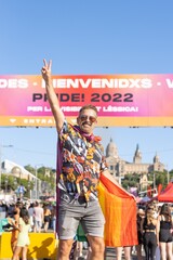 Happy smiling man on gay pride day in Barcelona with the pride flag, the background card which translated to English means Welcome to gay pride 2022.