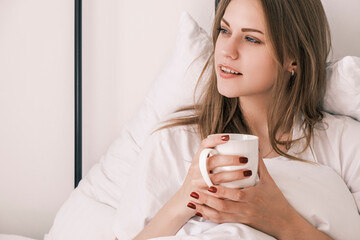 Fototapeta na wymiar Young attractive woman lies in bed after waking up with white cup in her hand looking out the window. Morning routine, time for yourself. Contemplation and enjoyment. Psychological self care
