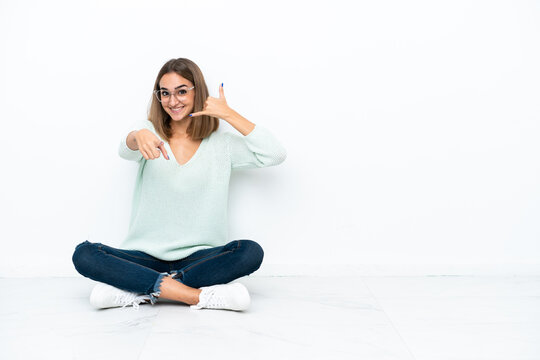 Young caucasian woman sitting on the floor isolated on white background making phone gesture and pointing front