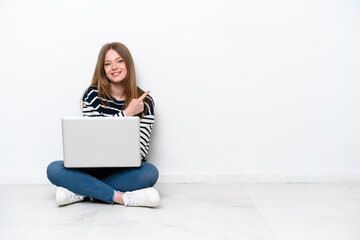 Young caucasian woman with a laptop sitting on the floor isolated on white background pointing to the side to present a product
