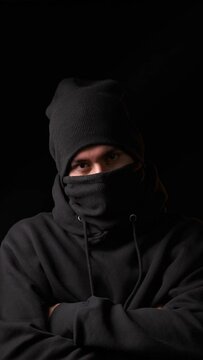 Thief in black balaclava and black cap staring at camera while standing against