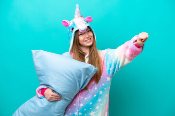 Young caucasian girl with unicorn pajamas holding pillow isolated on blue background giving a...