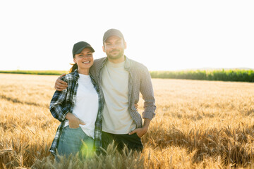 A couple of farmers in plaid shirts and caps stand embracing on agricultural field of wheat at...