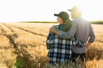 Naklejka na ściany i meble A couple of farmers in plaid shirts and caps stand embracing on agricultural field of wheat at sunset