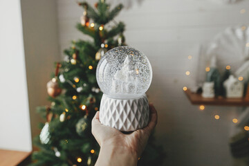 Stylish christmas snow globe in hand on background of christmas tree in lights in festive decorated...
