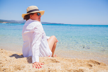 Fototapeta na wymiar Happy woman in white shirt and hat, relaxing on sand beach, enjoying the sea view. Summer travel lifestyle, holiday concept. Beautiful seascape of turquoise sea water. Back view. 