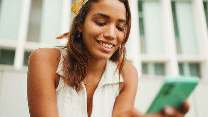 Profile of cute tanned woman with long brown hair wearing white top and yellow bandana seated using cellphone on bench in cityscape background.