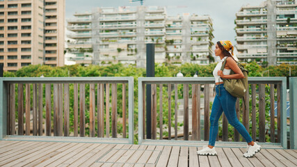 Сute tanned woman with long brown hair wearing white top and yellow bandana walks on bridge with backpack on her shoulder and looks at modern city