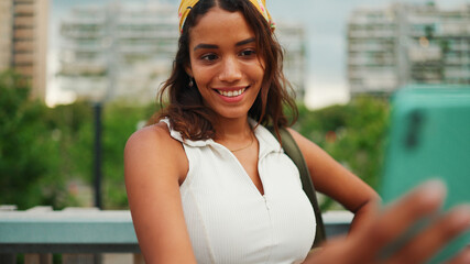 Cute tanned woman with long brown hair wearing white top and yellow bandana takes selfie while standing on bridge on modern city background