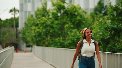 Cute tanned woman with long brown hair in white top and yellow bandana with backpack on her shoulders walks along the bridge.