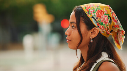 Profile of cute tanned woman with long brown hair in white top and yellow bandana with backpack on her shoulders is walking along pedestrian crossing