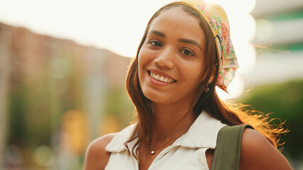 Clouse-up, cute tanned woman with long brown hair wearing white top and yellow bandana posing on camera. Smiling girl looking at the camera