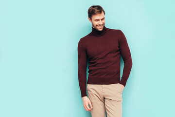 Portrait of handsome smiling stylish hipster lambersexual model. Sexy man dressed in turtleneck sweater and trousers. Fashion male isolated on blue wall in studio.