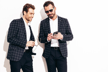 Two handsome confident stylish hipster lambersexual models. Sexy modern men dressed in black same elegant suit. Fashion male posing in studio near white wall. Shows something at his mobile phone