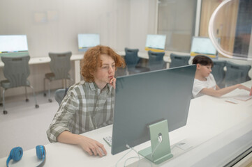 A ginger schoolboy sitting at the computer at school and looking involved