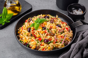 Close-up of Orzo pasta salad with vegetables, olive taggiasche, tomato, bell pepper, chickpeas, red onion, basil. Olive oil, Dark gray table surface.