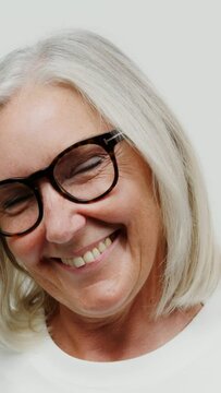 An adult gray-haired woman with glasses smiles, grimaces while looking at camera