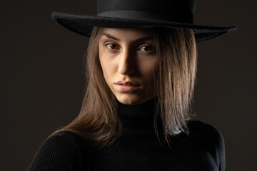 Beautiful woman. Close-up of girls face. Long-haired woman in hat. Brown-haired business lady looks at camera. Serious girl on dark background. Attractive woman model. Female student in black clothes