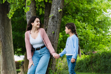 young woman and child holding hands and looking at each other near trees in park