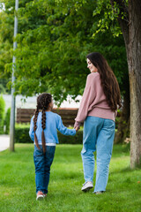Fototapeta premium back view of mother and daughter in jeans holding hands while walking in park