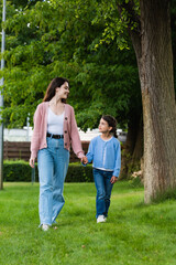 happy woman and girl holding hands and looking at each other while walking in park