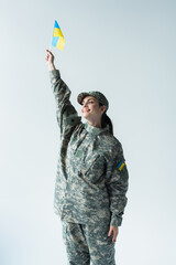 Smiling soldier in uniform rising ukrainian flag isolated on grey