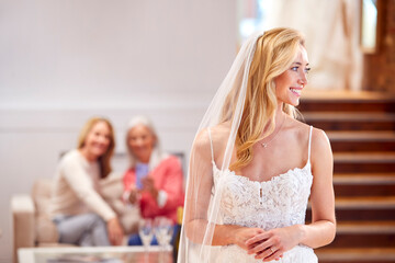 Grandmother With Mother Watching Adult Daughter Choosing And Trying On Wedding Dress In Bridal Store