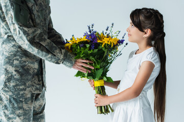 Child giving blue and yellow bouquet to servicewoman in camouflage uniform isolated on grey